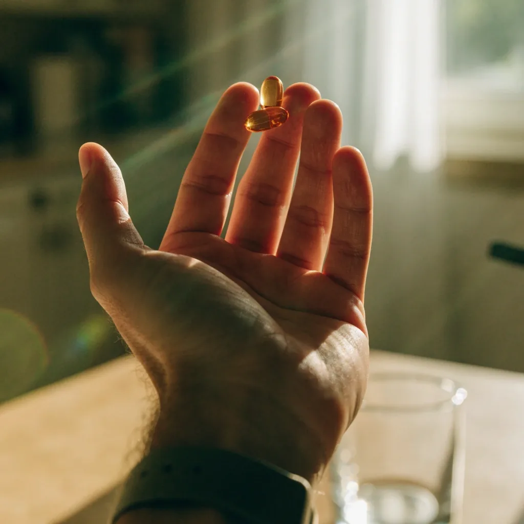 Open hand holding two softgel capsules in front of a soft-lit kitchen counter with a water glass nearby